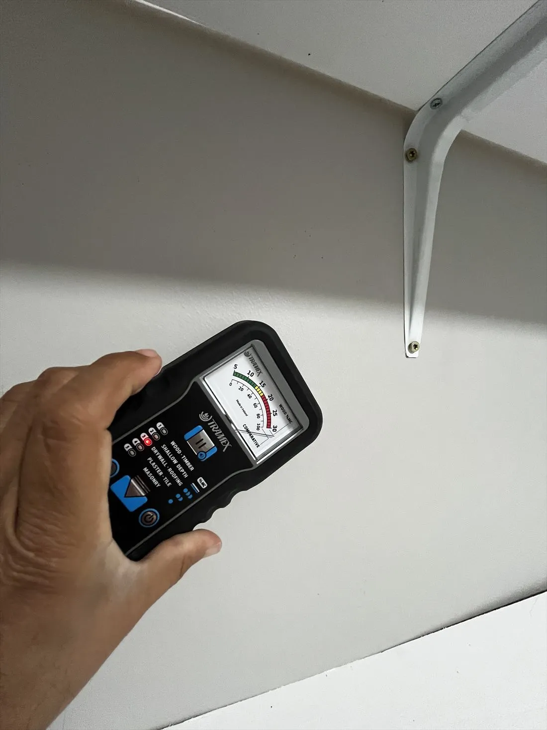Hand holding a moisture meter against a wall below a shelf bracket